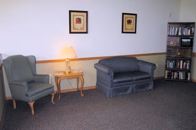 A small sitting area with a gray upholstered armchair on the left, a wooden side table with a lamp in the center, and a blue patterned loveseat on the right. Above the loveseat are two framed floral pictures. To the far right is a wooden bookshelf filled with books and some decorative items. The room has beige walls with a wooden chair rail and carpeted floor.
