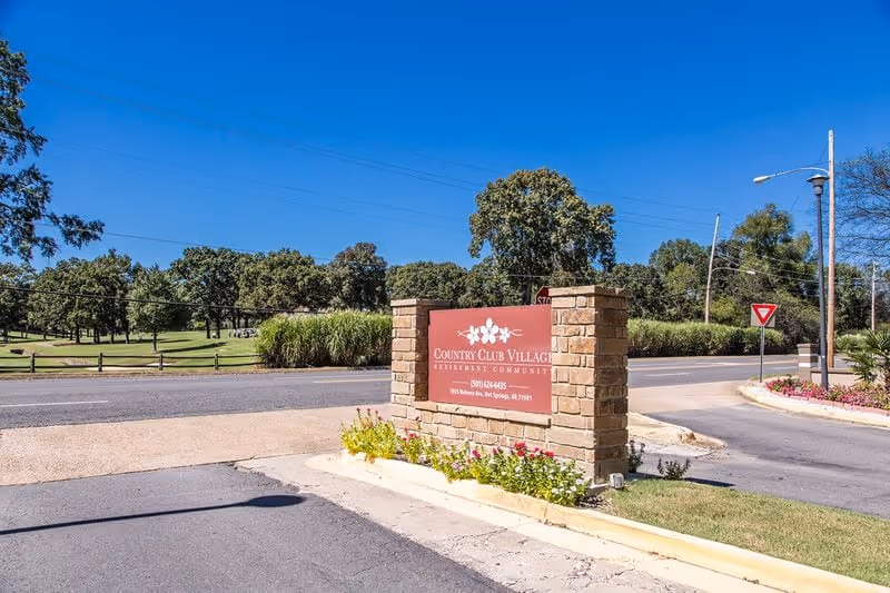 Entrance sign for Country Club Village Retirement Community made of brick with a red plaque displaying the community name and contact information, surrounded by flowers and greenery, with a road and trees in the background under a clear blue sky.