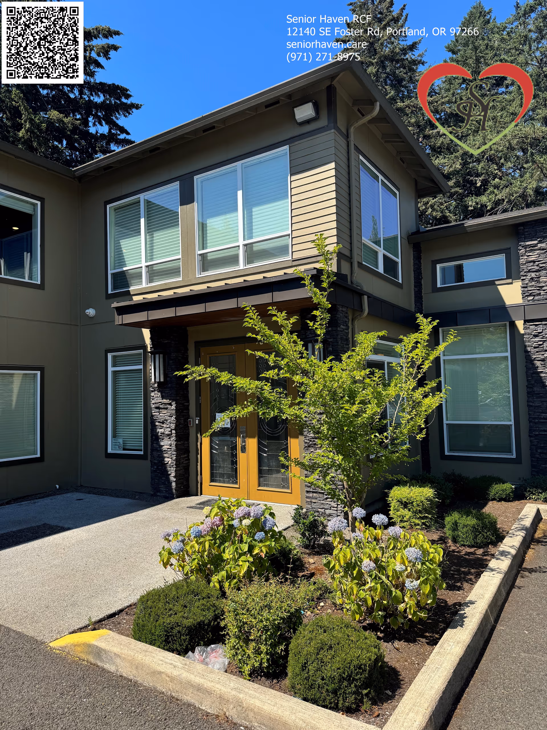 Exterior view of Senior Haven RCF building entrance with large windows, a double wooden door, and landscaped garden featuring bushes and flowering plants under a clear blue sky.