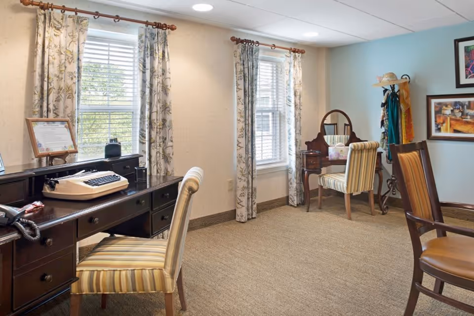 A well-lit room with two windows covered by floral curtains. There is a dark wooden desk with a vintage typewriter and a telephone, accompanied by a striped upholstered chair. In the corner, there is a wooden vanity with a mirror and another striped chair. A coat rack with hats and scarves stands nearby, and framed artwork hangs on the light blue wall.