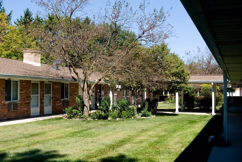 Outdoor view of a senior living facility with a grassy courtyard, trees, and a brick building with multiple doors and windows. There is a covered walkway on the right side and some outdoor chairs near the building.