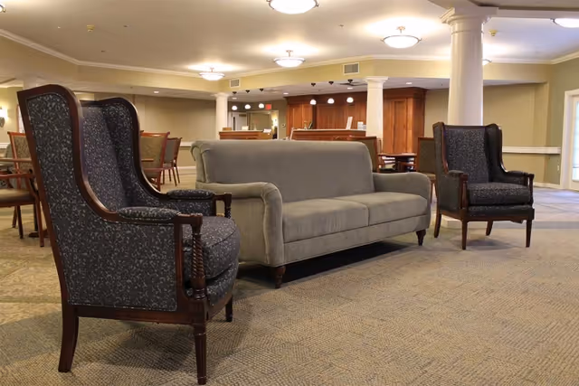 Open communal lounge with a gray sofa, two upholstered wingback chairs, dining tables and a serving counter in the background.