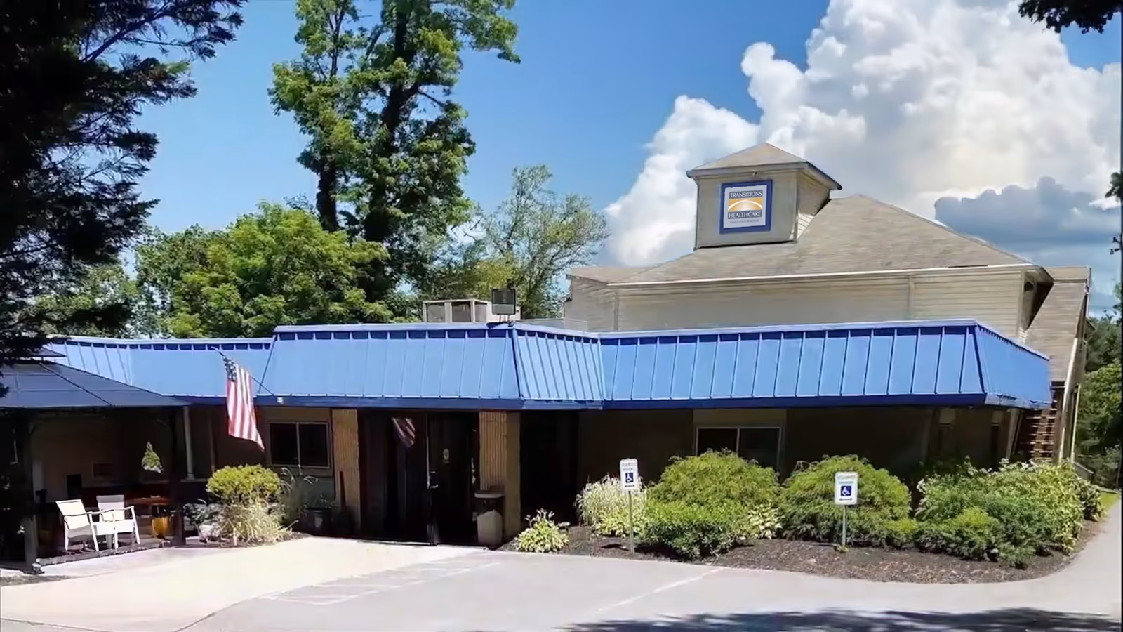 Exterior view of Transitions Healthcare Oakland Manor building with a blue awning, American flag, and surrounding greenery under a partly cloudy sky.