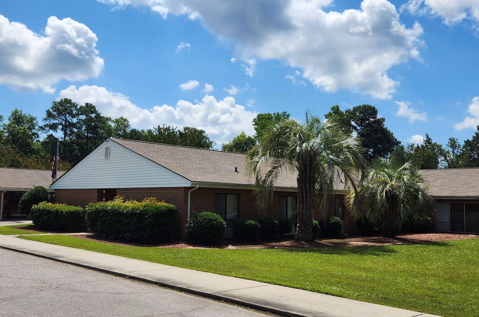 Single-story brick senior care facility exterior with manicured lawn and palm trees against a blue sky.