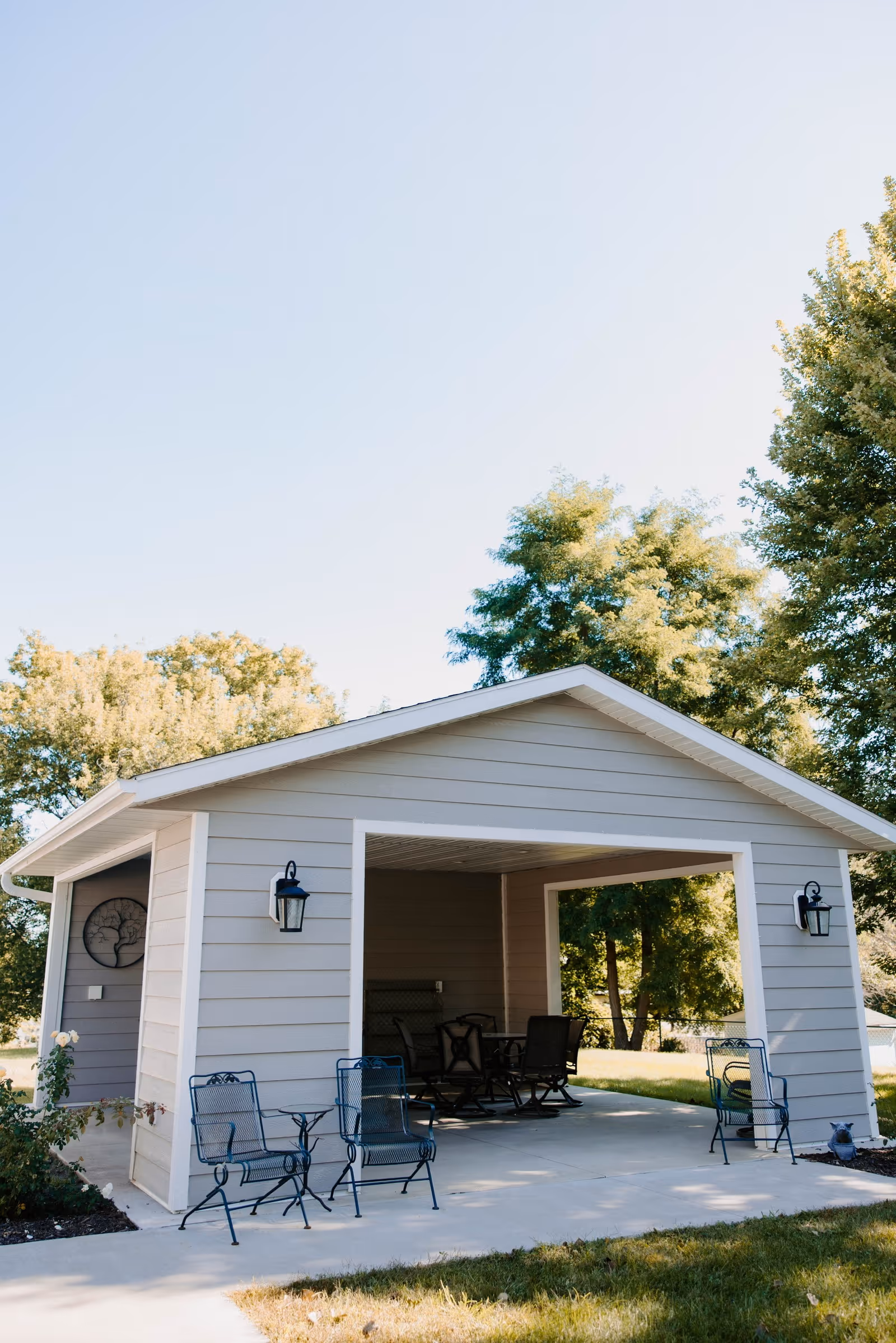 Open-sided light-gray outdoor pavilion with patio furniture on a concrete slab surrounded by trees and lawn.