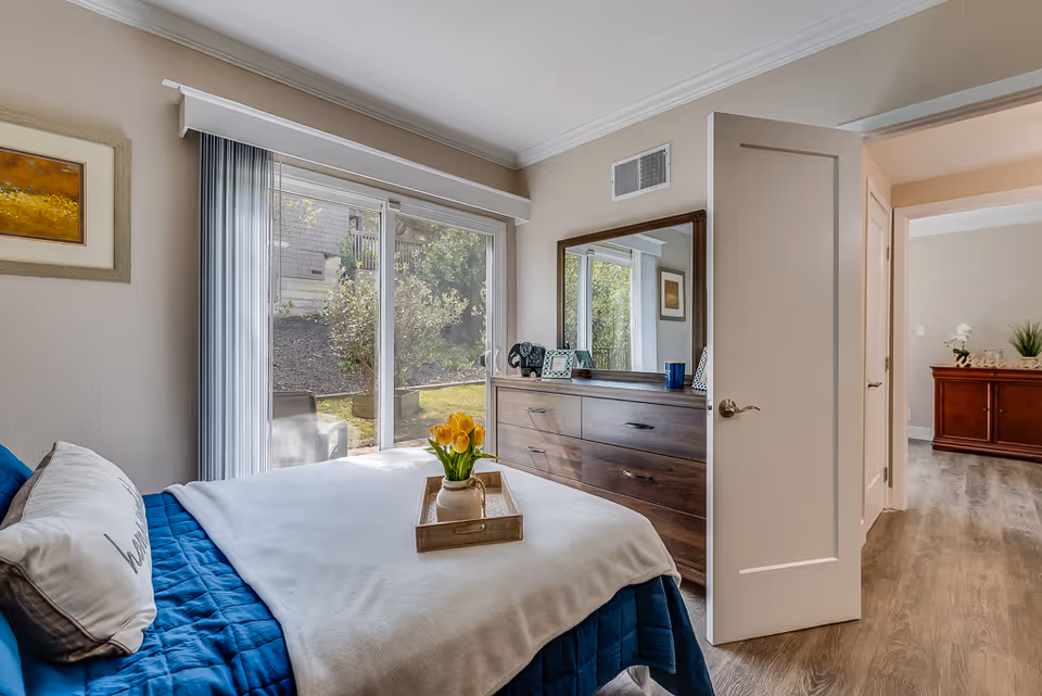 Sunlit bedroom with blue bedding, a tray of yellow tulips on the bed, sliding glass doors to a patio, and a wooden dresser with mirror next to an open door.
