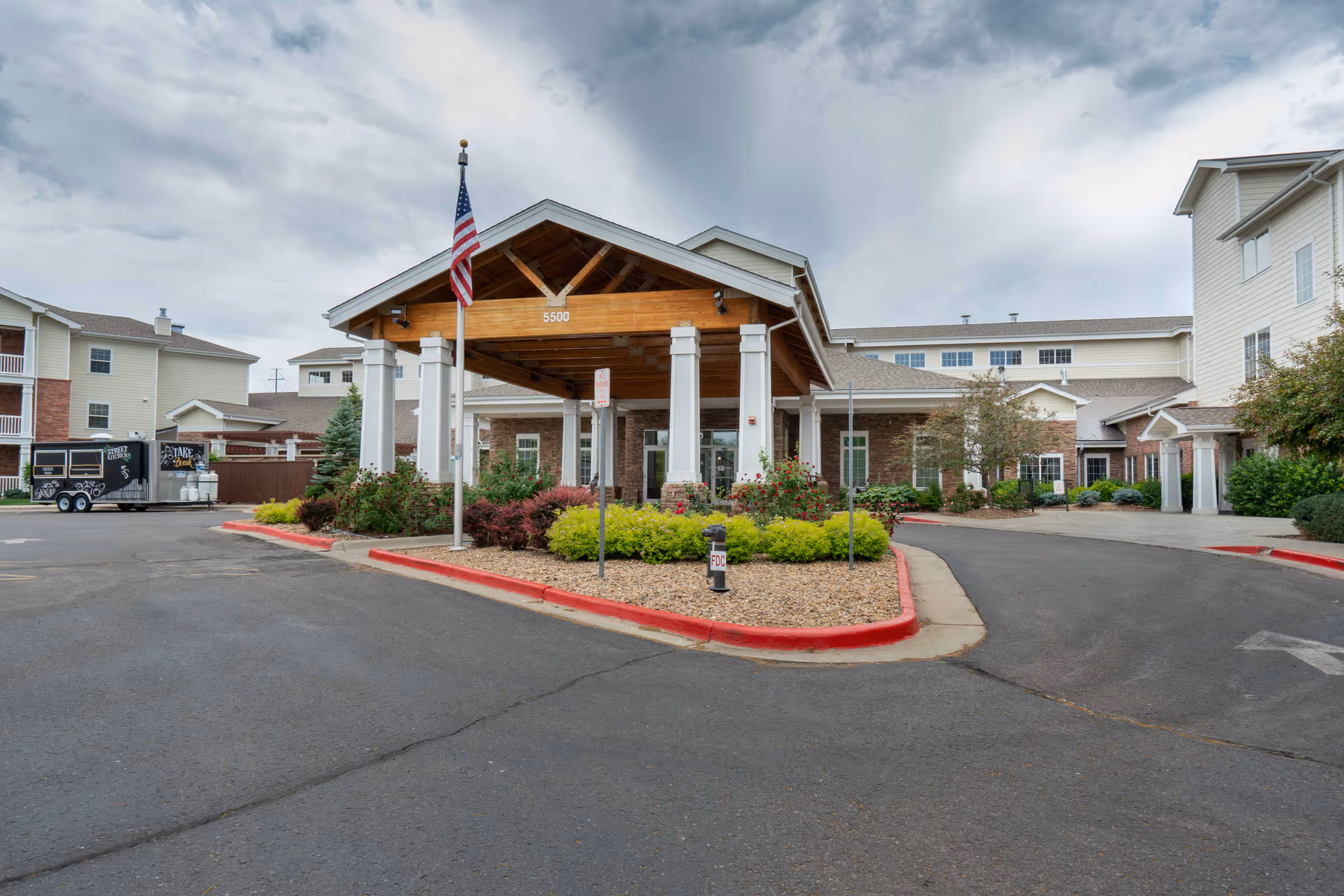 Entrance of a senior living facility named Holly Creek with a covered drop-off area supported by white columns and a wooden roof. The building is surrounded by landscaped bushes and plants, with an American flag flying on a pole in front. The sky is cloudy and the driveway curves around the entrance.