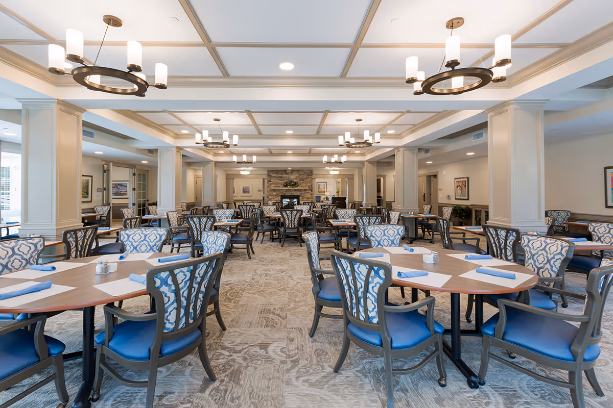 A spacious dining room in an assisted living facility with multiple round tables and chairs arranged neatly. The chairs have blue cushioned seats and patterned backs. The room features a coffered ceiling with modern chandeliers, beige walls with white trim, and a carpeted floor with a subtle pattern. In the background, there is a stone fireplace with artwork on the walls.