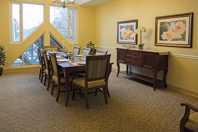 A dining room with a long wooden table set for eight people with plates, glasses, and silverware. The room has yellow walls, a patterned carpet, and a large window with a triangular design letting in natural light. There is a wooden sideboard against the wall with two framed floral paintings above it and a small plant on top.