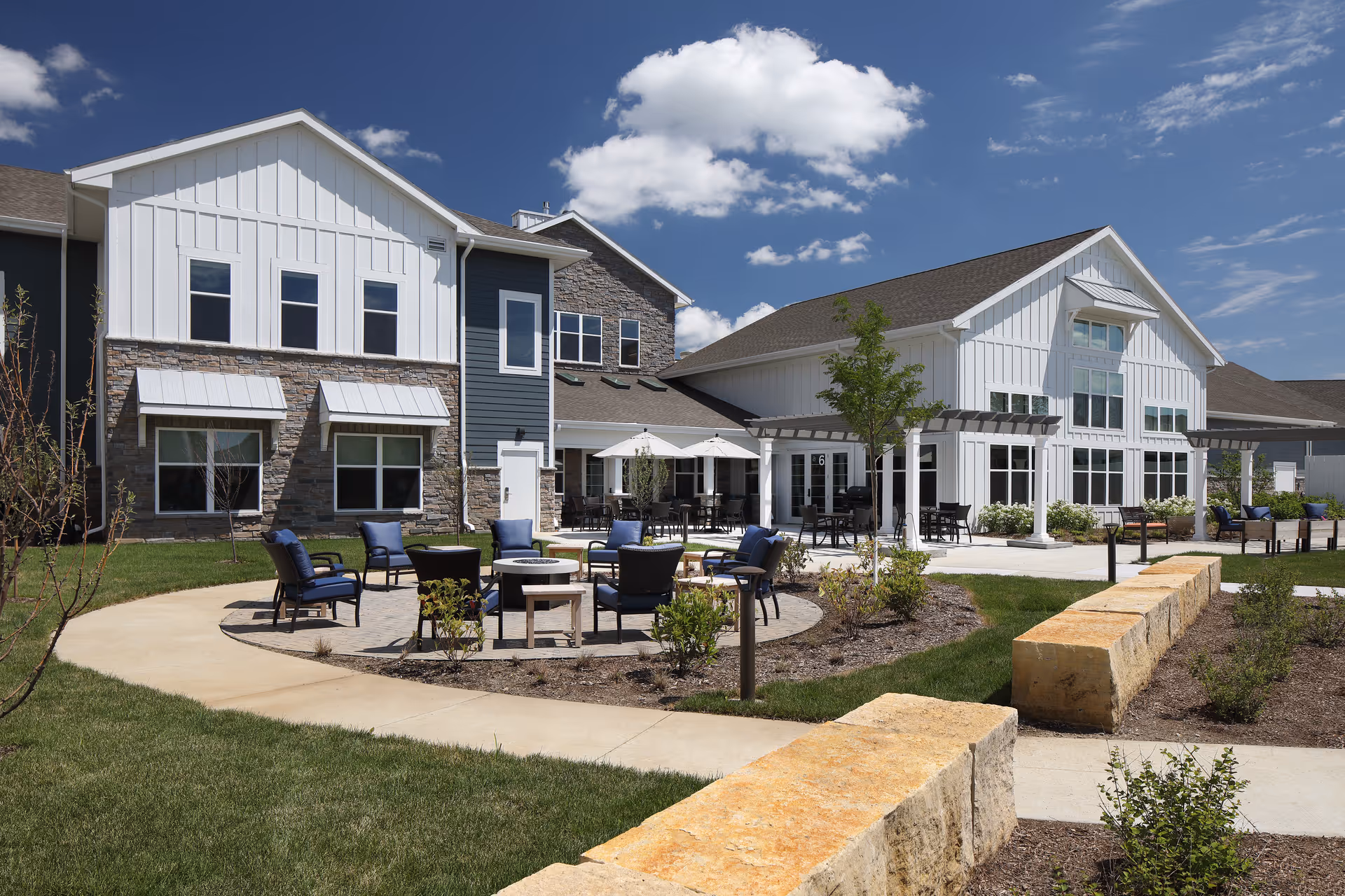 Outdoor seating area and landscaped courtyard in front of a modern senior living facility building under a blue sky.