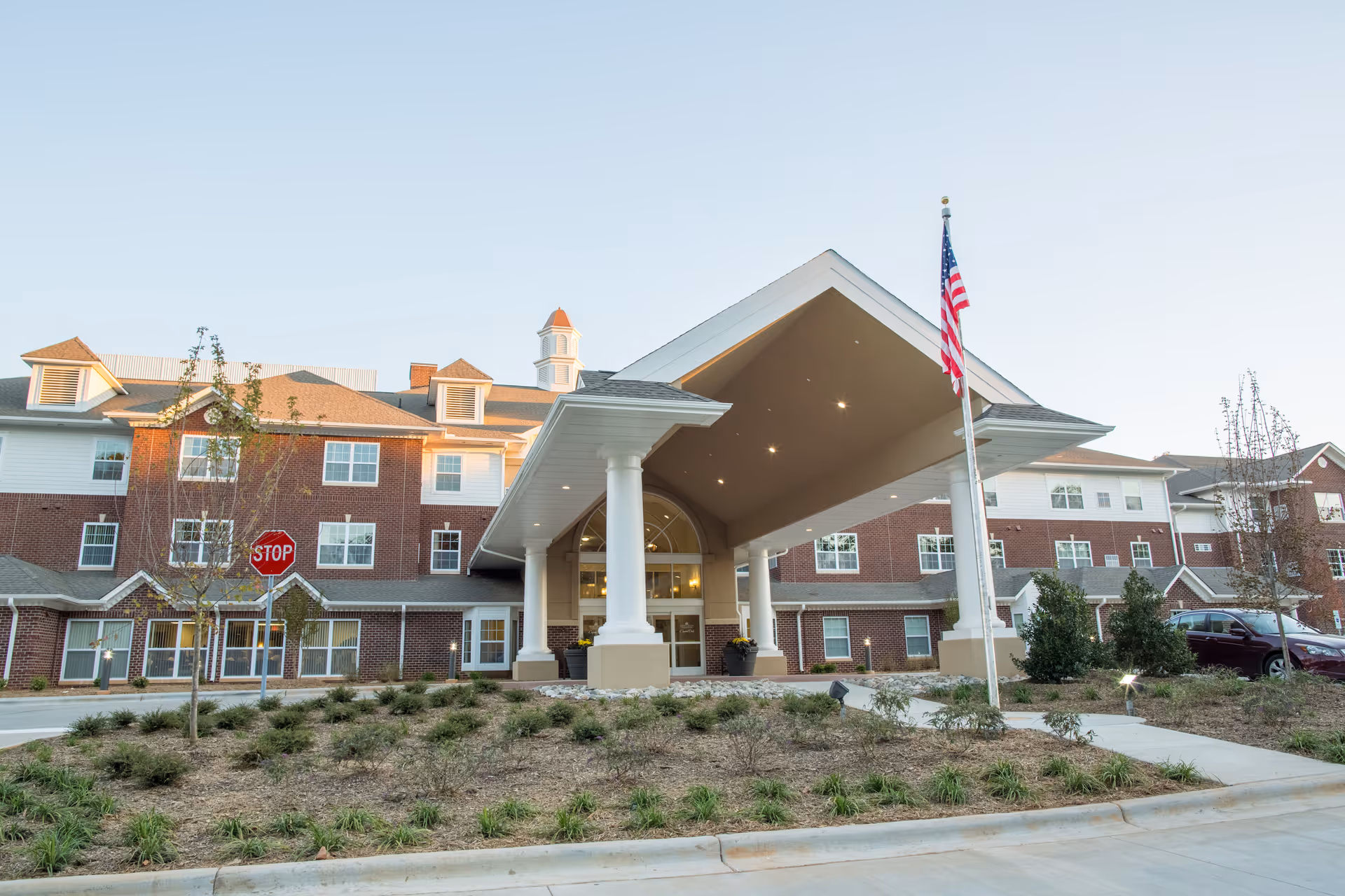 The front entrance of a brick senior living building with a covered porte-cochère, American flag, and landscaped driveway.