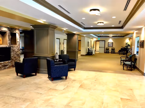 Spacious senior living lobby with seating areas, decorative columns, a stone fireplace, and a piano in the background.