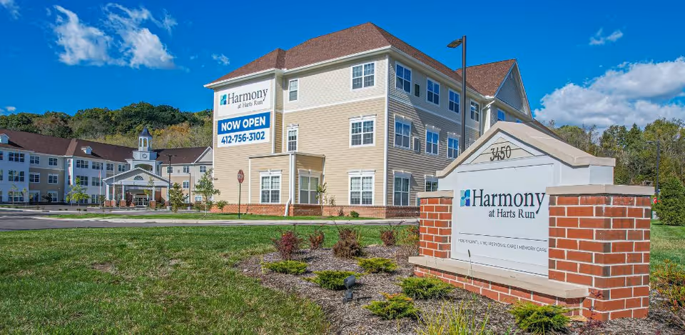 Exterior view of Harmony at Harts Run senior living facility with a large brick sign in the foreground displaying the facility name and address number 3450. The building is beige with white trim and has multiple windows. A sign on the building indicates it is now open with a contact phone number. The sky is blue with some clouds and there is greenery and landscaping around the property.