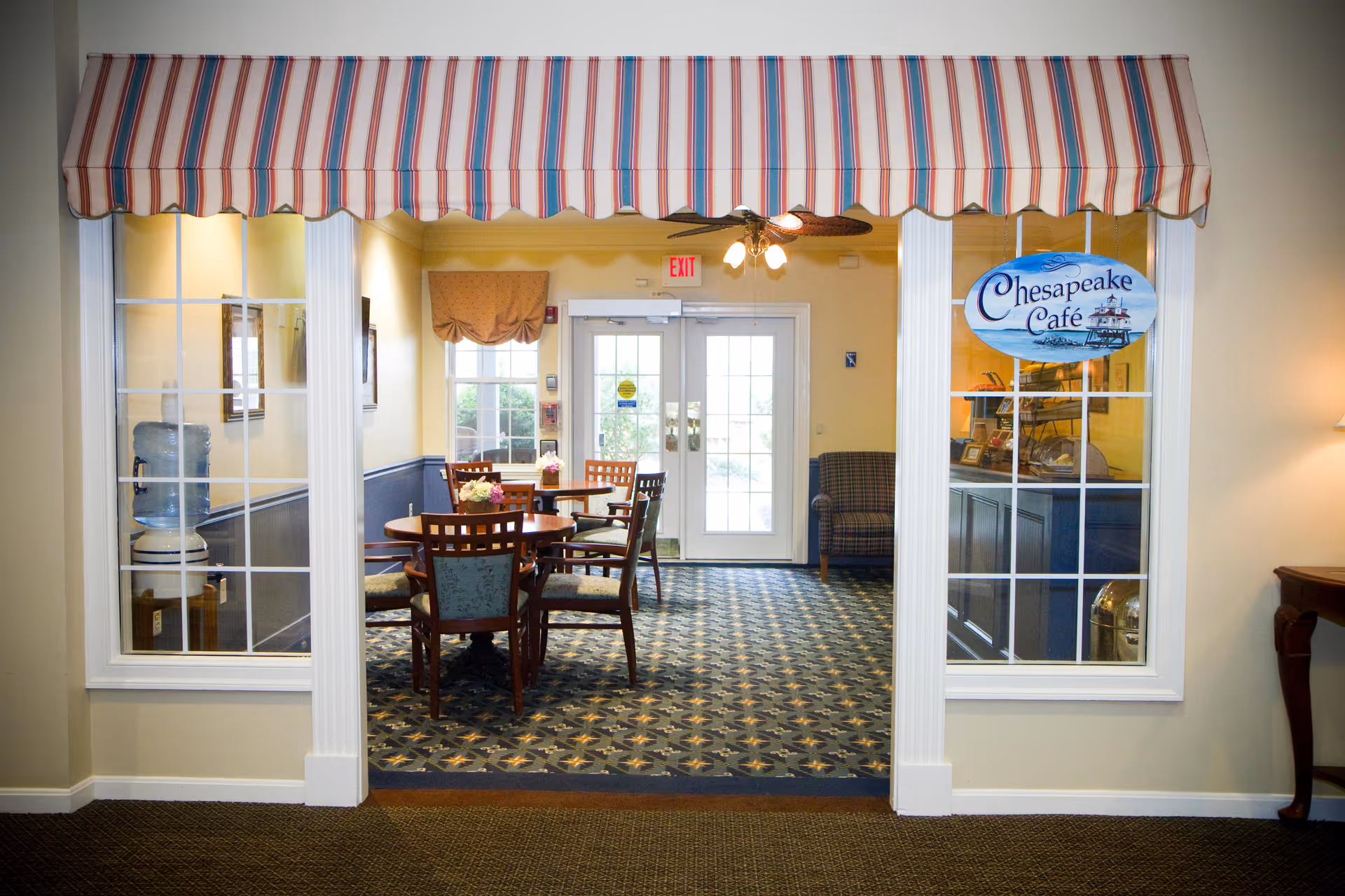 Entrance to Chesapeake Café with a striped awning above. Inside, there are round tables with chairs, a water cooler on the left, and a plaid armchair near the back door. The room has patterned carpet and yellow walls with white trim.