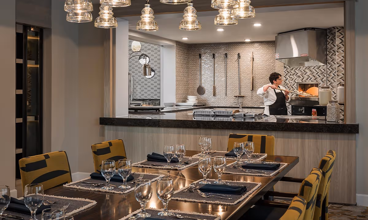 A dining room with a set table and place settings in the foreground and an open kitchen where a chef is preparing food in the background.