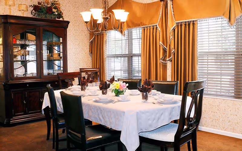 A formal dining room with a rectangular table covered with a white tablecloth, set with white dishes, cups, and brown folded napkins. There are six dark wooden chairs around the table. A chandelier with six lights hangs above the table. Behind the table is a large window with gold curtains and blinds. To the left is a dark wooden china cabinet with glass doors displaying dishes and decorative items.