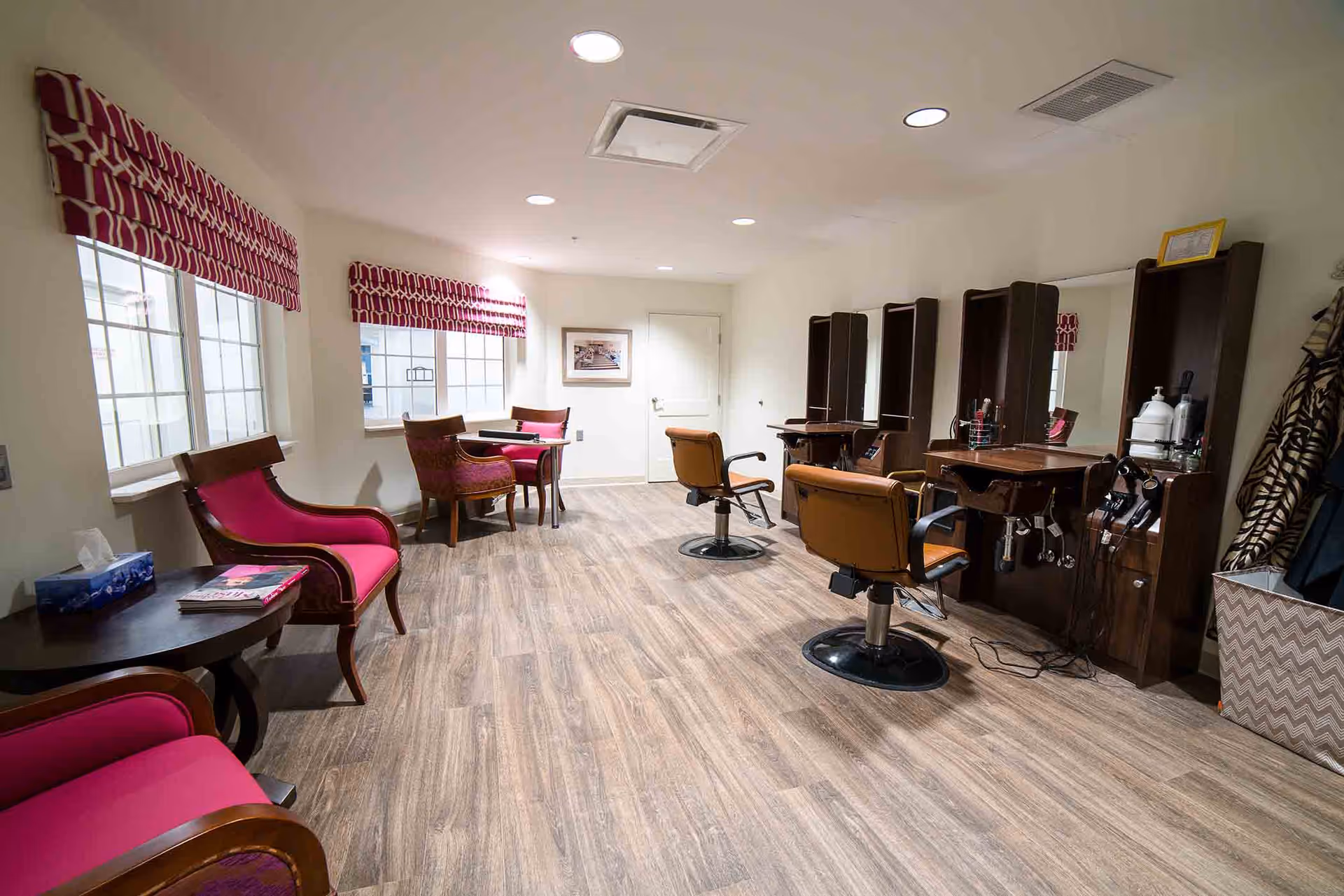 Interior view of a salon area in a senior living facility with wooden flooring, three salon chairs in front of mirrors and styling stations on the right, and several upholstered chairs with pink cushions and small tables near windows with red patterned valances on the left.
