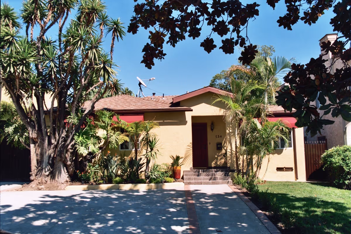 Front exterior view of a single-story house with a brown roof and beige walls, surrounded by various palm trees and plants. The driveway leads up to the front door with steps, and there is a satellite dish on the roof. The sky is clear and blue.