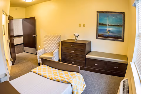 A small, cozy bedroom with a single bed covered with a white sheet and a yellow patterned blanket. There is a cushioned armchair next to a dark wooden dresser with a small vase of flowers on top. Next to the dresser is a cushioned bench with storage drawers underneath. A framed painting of a sailboat on water hangs on the wall above the bench. The room has beige walls, a window with blinds, and an open door leading to a carpeted hallway.
