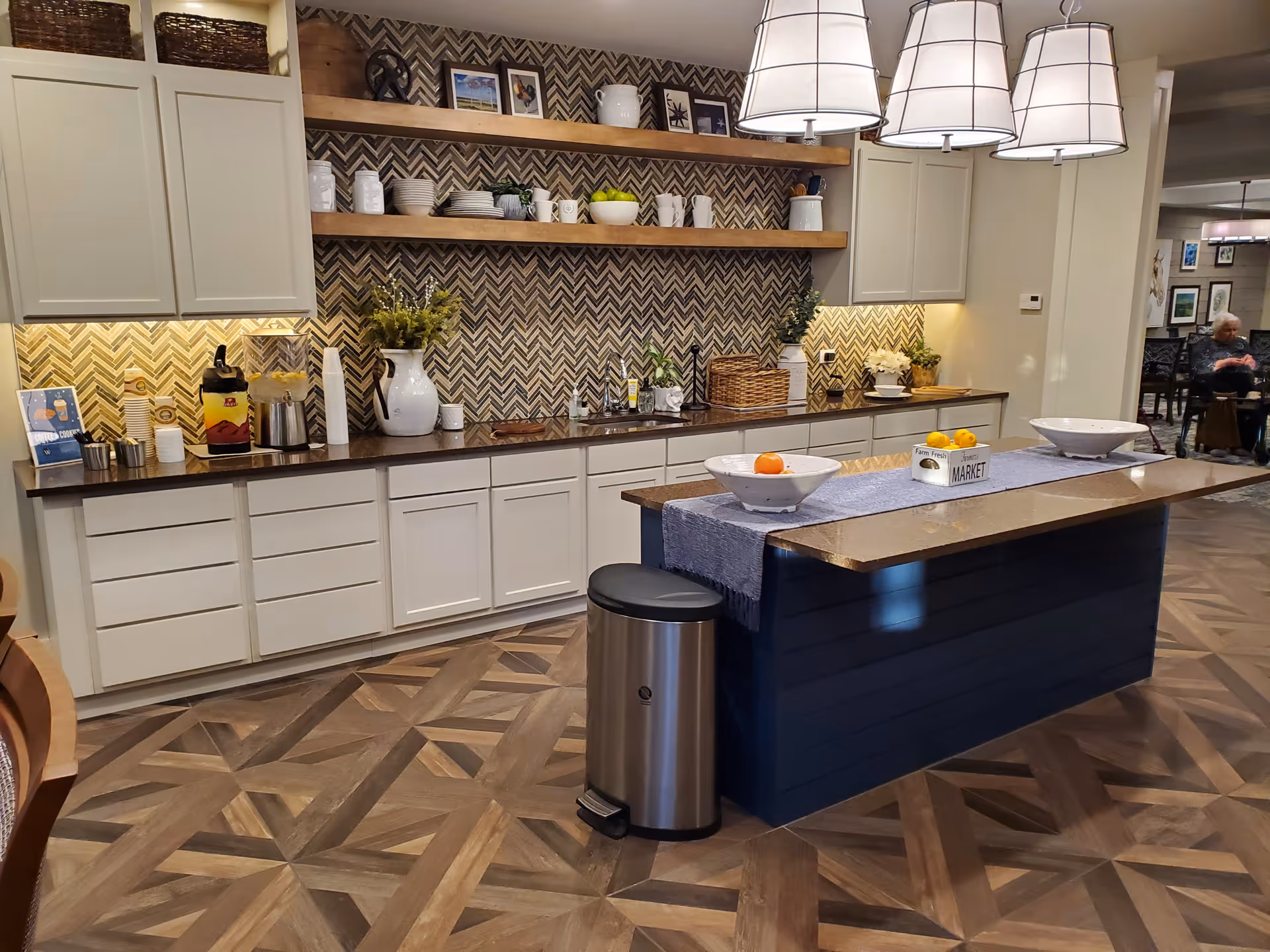 A modern kitchen area with white cabinets, dark countertops, and a patterned backsplash. There are two wooden shelves holding dishes and decorative items. A kitchen island with a blue base and a granite countertop is in the center, adorned with a blue runner and bowls of fruit. Three large pendant lights hang above the island. A stainless steel trash can is next to the island. In the background, a person is seated in a dining or common area.