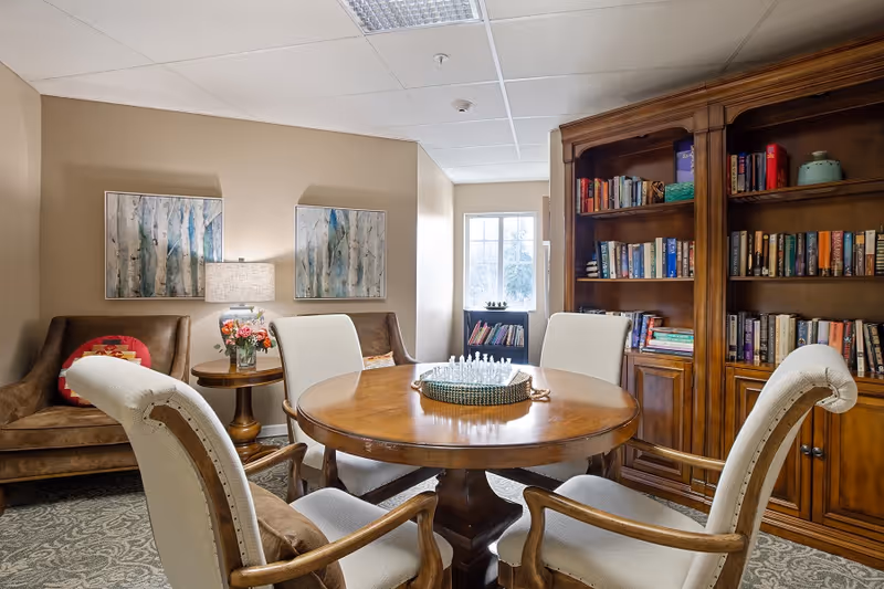A cozy room with a round wooden table surrounded by four upholstered chairs. On the table is a chess set. Behind the table is a large wooden bookshelf filled with books. To the left, there are two brown armchairs with a small round table between them, holding a lamp and a vase of flowers. Two framed artworks hang on the beige wall above the armchairs. A window lets in natural light in the background.