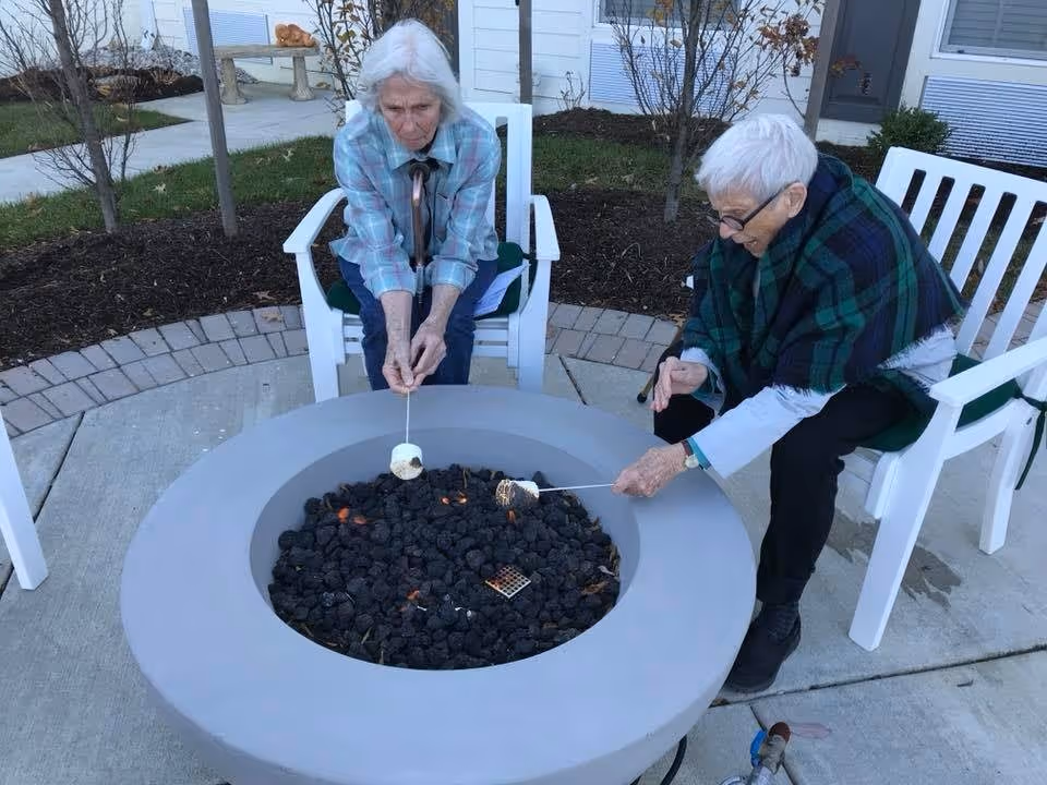 Two elderly women sitting on white chairs outdoors, roasting marshmallows over a circular fire pit filled with black lava rocks. One woman wears a plaid shirt and the other has a green and blue shawl. They are on a concrete patio with some landscaping and a white building in the background.