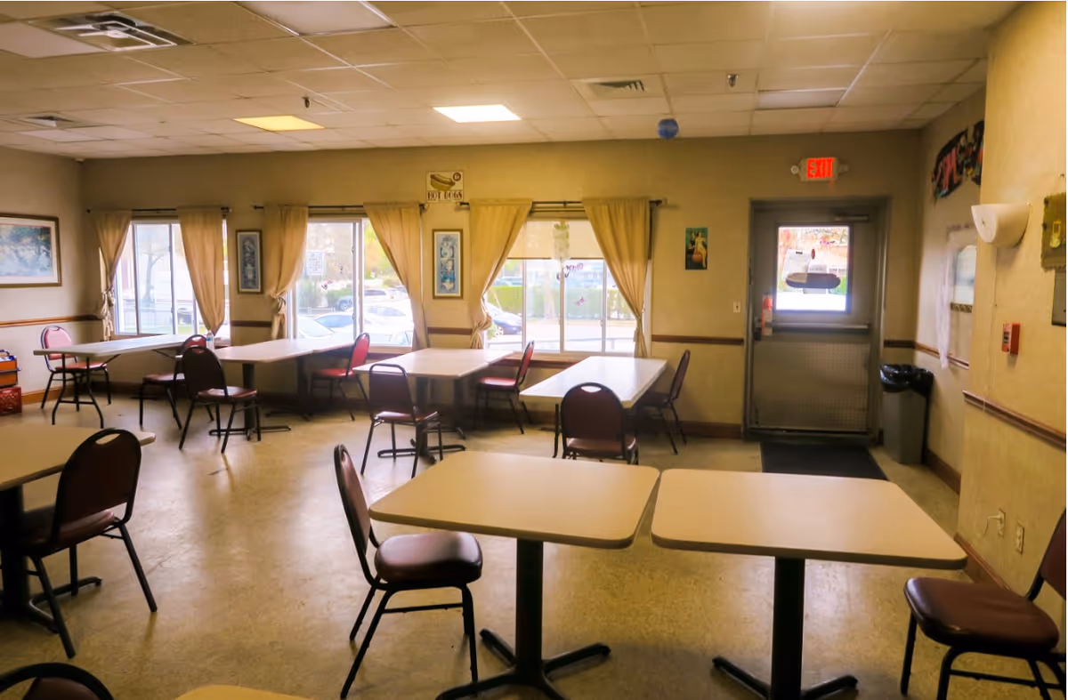 A dining room with several tables and chairs arranged neatly. The room has large windows with beige curtains allowing natural light to enter. There are framed pictures on the walls and an exit door at the far end. The floor is a light-colored linoleum, and the ceiling has recessed lighting panels.