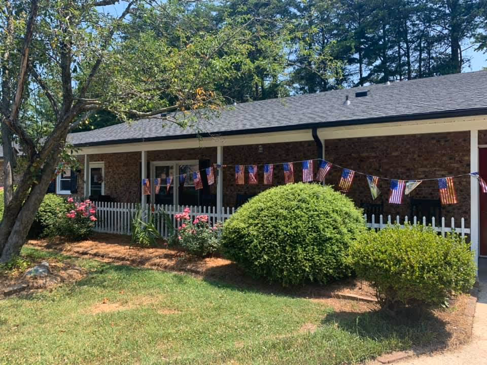 Exterior view of a single-story brick building with a gray roof, white picket fence, and a garden with bushes and flowers. American flag-themed pennant banners are strung along the fence. Trees and a clear blue sky are visible in the background.