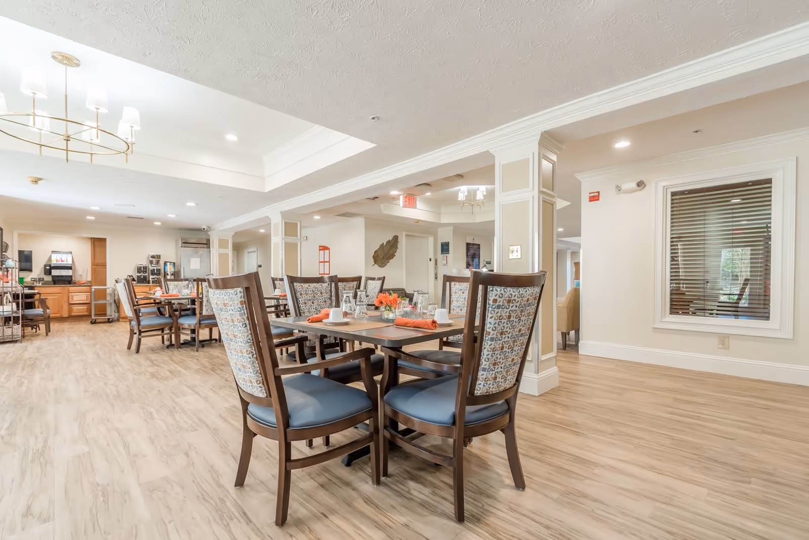 A bright and spacious dining room in a senior living facility with wooden tables and chairs arranged neatly. The tables are set with cups, glasses, and orange napkins. The room features light-colored walls, wood flooring, and a ceiling light fixture. In the background, there is a beverage station with coffee machines and cabinets.