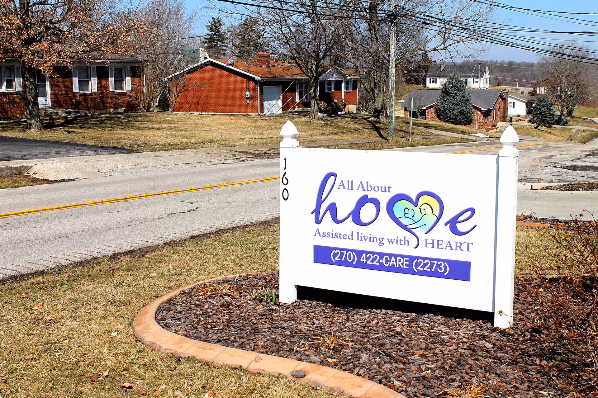 A white sign with purple and blue text reading 'All About hope Assisted living with HEART' and a phone number (270) 422-CARE (2273) is placed on a mulched area next to a road. Behind the sign, there are several single-story brick buildings and leafless trees under a clear sky.