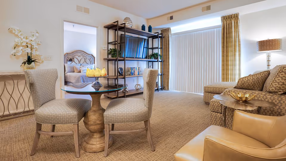 Bright furnished living room with a small round table and chairs, sofa and shelving unit, and a bedroom visible through an open doorway.