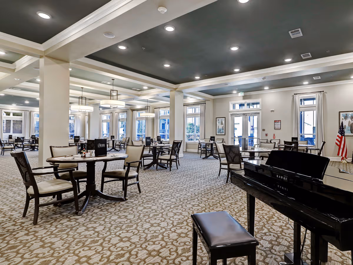 Spacious dining room with multiple round tables and chairs arranged neatly. The room has large windows with curtains, a patterned carpet, and a black grand piano with a bench in the foreground. Ceiling lights illuminate the space, and an American flag is visible near the back wall.