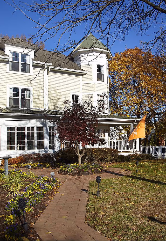 Exterior view of a multi-story senior living facility building with beige siding, multiple windows, and a small tower-like structure. A brick pathway leads through a landscaped garden with flowers and small trees. An orange flag with the word 'Sunrise' is visible near the porch. Trees with autumn foliage surround the building under a clear blue sky.