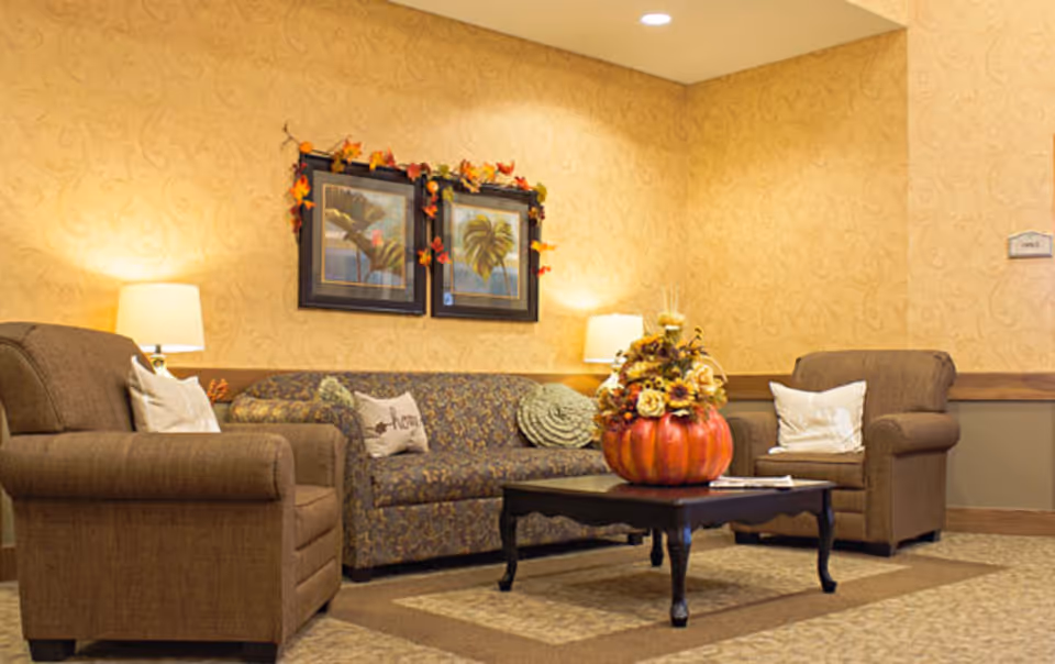 A cozy senior living facility lounge area with two brown armchairs and a patterned sofa. The sofa has decorative pillows, and a coffee table in front holds a large pumpkin-shaped floral arrangement. Two framed pictures with autumn leaves garland hang on the beige patterned wall, and two table lamps provide warm lighting.