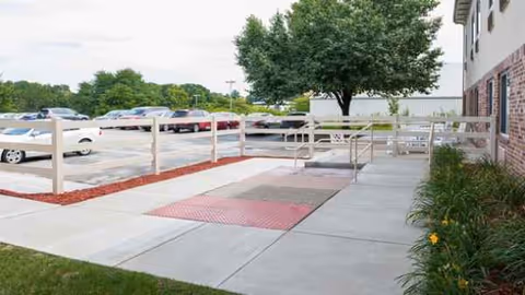 Exterior view of Monroeville Skilled Nursing and Rehabilitation Center with walkway, fence, parking lot, and trees.