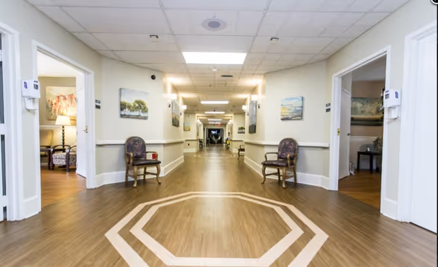 A long, well-lit hallway in a senior living facility with wooden flooring and a geometric pattern near the foreground. The hallway has chairs placed along the walls and several paintings hanging. Open doorways on either side reveal rooms with comfortable seating and lamps.
