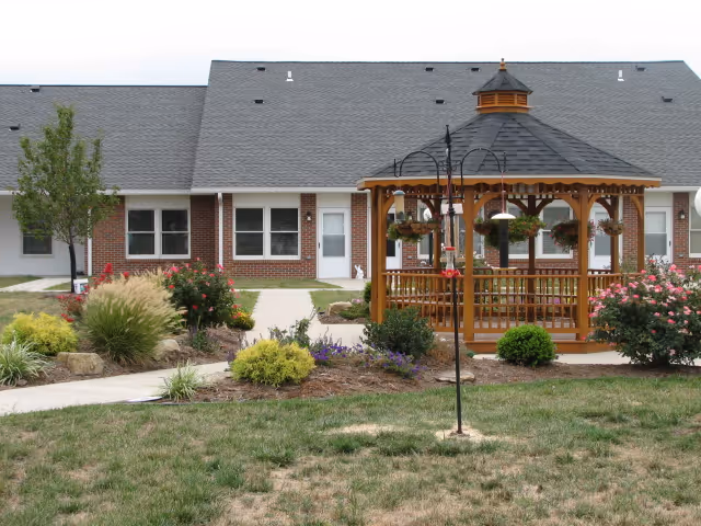 A landscaped outdoor area in front of a single-story brick building with white trim and a gray roof. The area features a wooden gazebo with hanging flower baskets, surrounded by various shrubs, flowers, and a small tree. A paved walkway leads to the building entrance and around the gazebo.