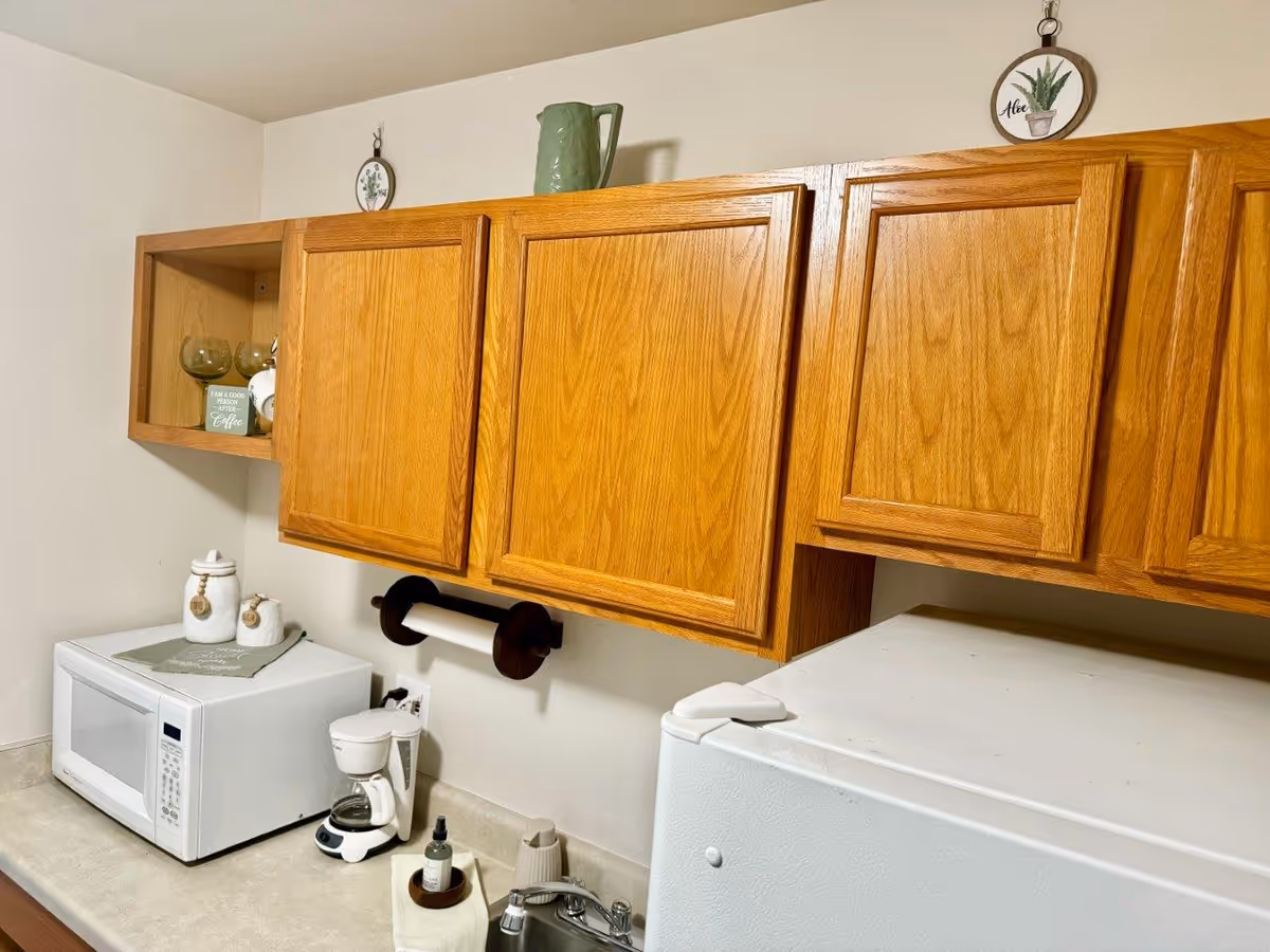 Small kitchenette with oak wall cabinets above a microwave, coffee maker, sink and a compact refrigerator.