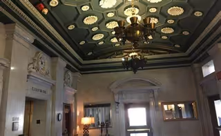 Interior view of an elegant hallway or lobby area with ornate ceiling decorations, a large chandelier, marble walls, and a lamp on a table near the entrance door.