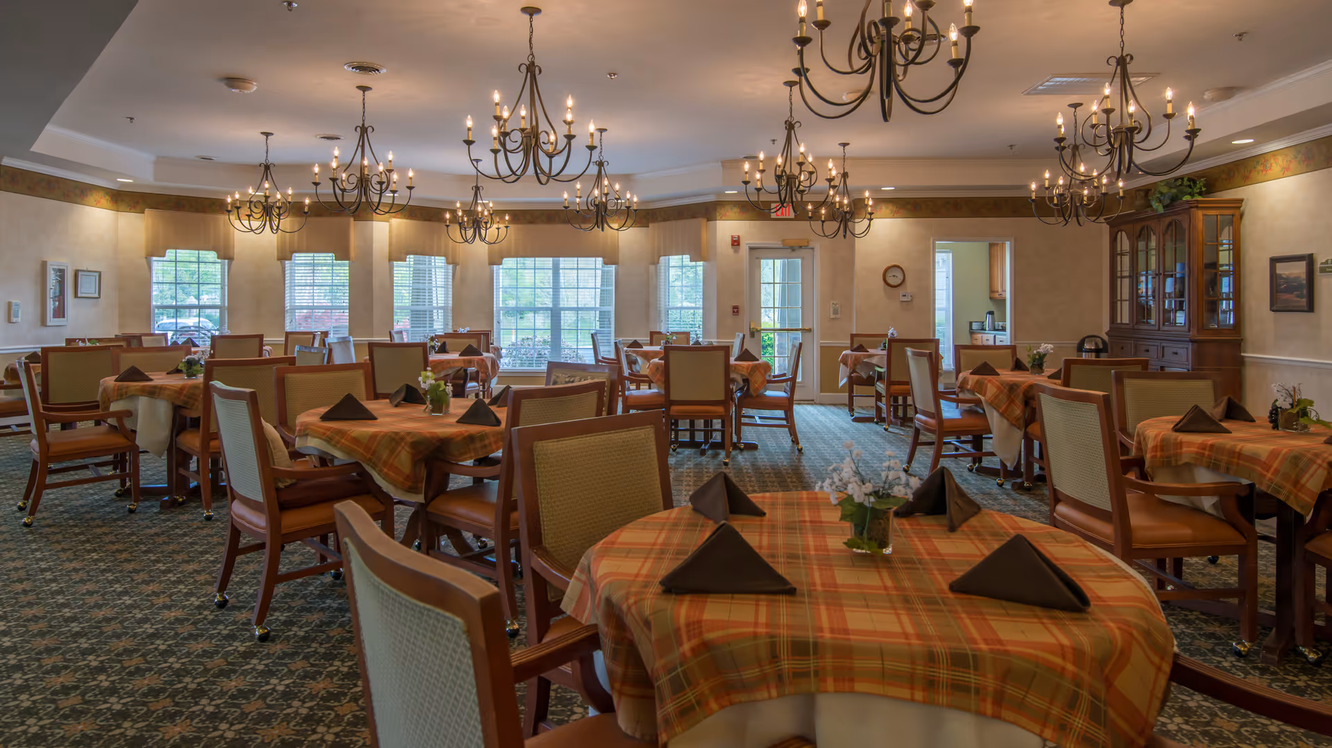 A spacious dining room with multiple tables covered in orange plaid tablecloths, each set with folded dark napkins and small floral centerpieces. The room features several large windows letting in natural light, elegant chandeliers hanging from the ceiling, and wooden chairs with cushioned seats. There is a wooden cabinet on the right side and a door leading to another room in the background.