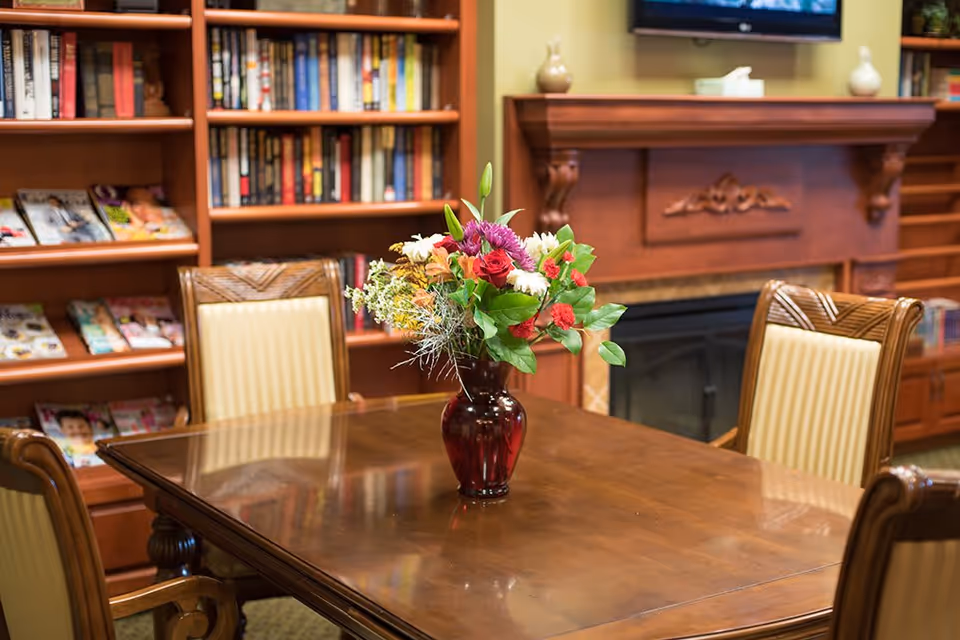 A wooden table with a vase of colorful flowers in the center, surrounded by four wooden chairs with beige cushioned seats. In the background, there is a wooden bookshelf filled with books and magazines, and a fireplace with a wooden mantel and a TV mounted above it.