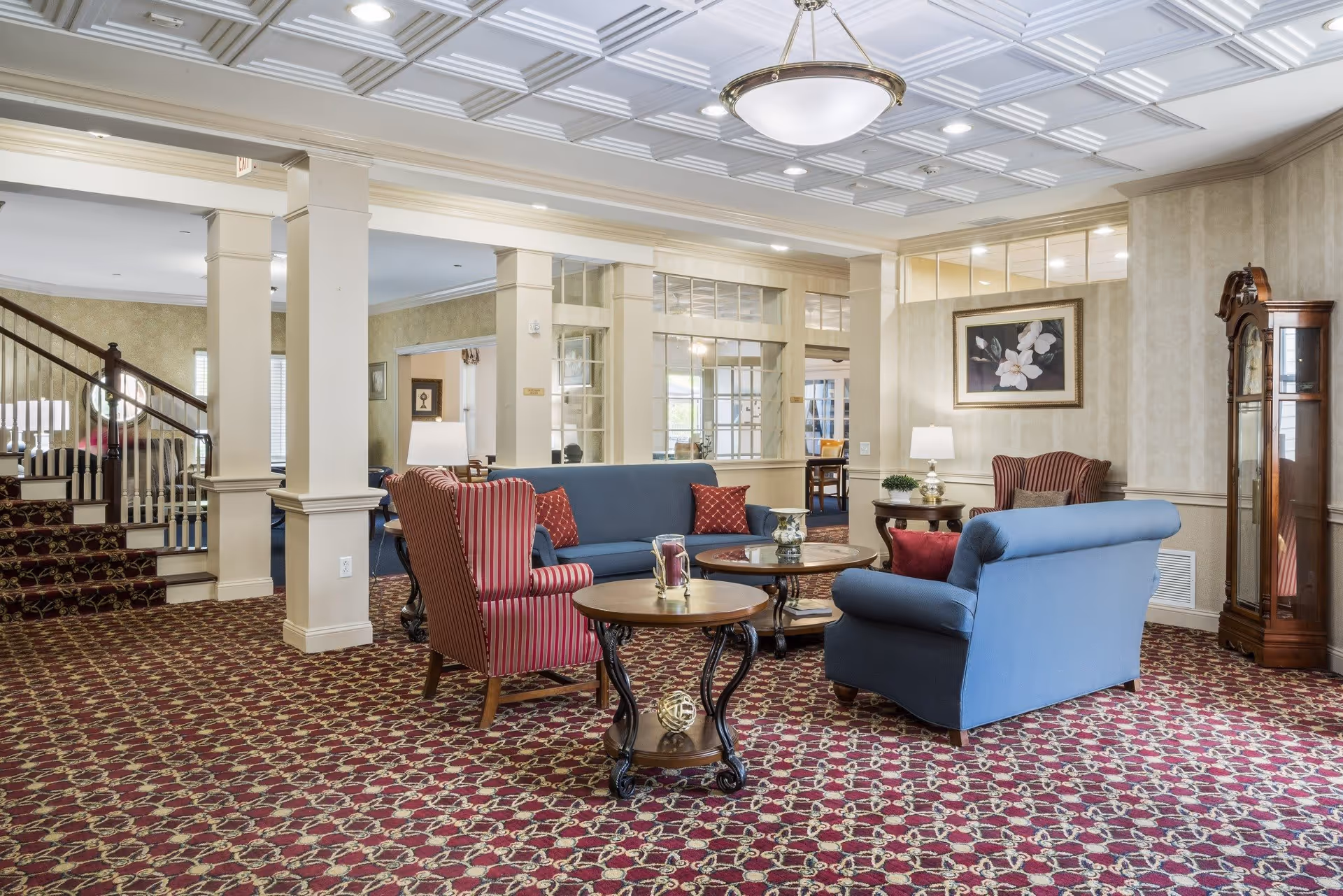 A cozy senior living facility lounge area with patterned red carpet, blue sofas, and red striped armchairs arranged around wooden coffee tables. The room features cream-colored walls, a grandfather clock, a floral painting, and a staircase with a dark wooden banister. Large windows and glass panels allow natural light to brighten the space.