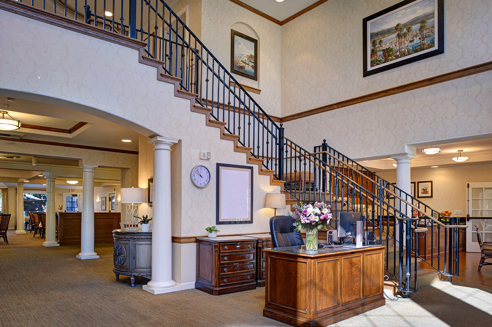 Reception desk with fresh flowers beneath an elegant staircase in a senior living facility lobby.