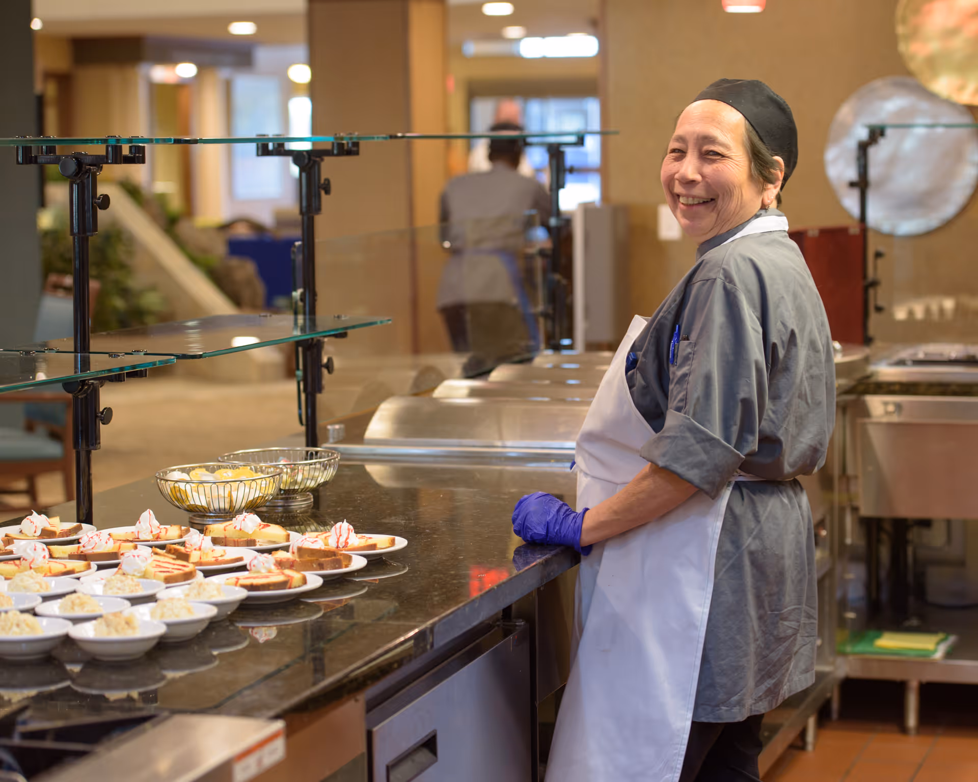 A smiling kitchen staff member wearing a gray uniform, white apron, black hairnet, and purple gloves stands behind a counter with plates of desserts in a commercial kitchen setting.