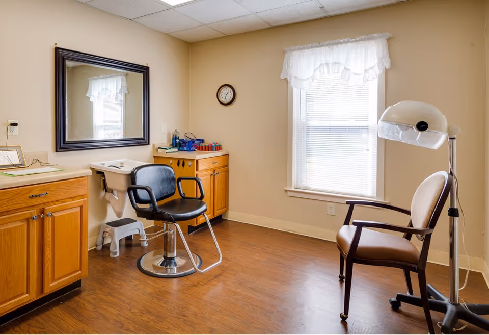Interior of a small hair salon or grooming room with a black salon chair in front of a sink, wooden cabinets, a large mirror on the wall, a clock, a window with white curtains, and a beige armchair next to a hair dryer.
