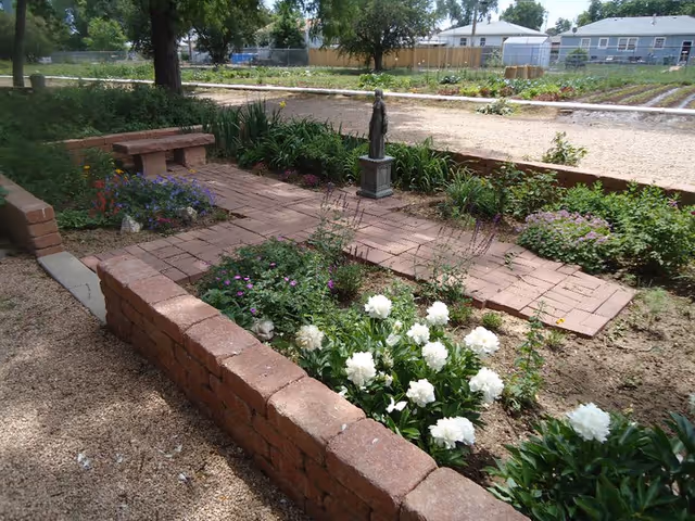 A peaceful outdoor garden area with brick pathways and raised flower beds containing various plants and white flowers. There is a small statue on a pedestal in the center of the garden, surrounded by greenery. In the background, there are houses and a fenced area with more plants.