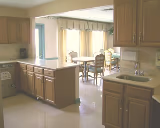 Bright kitchen with wood cabinets, a sink and a countertop island opening onto a dining area with tables and chairs.