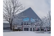 Exterior view of a building with a steep triangular roof covered in snow. Snow-covered trees and ground are visible in the foreground under a cloudy sky.