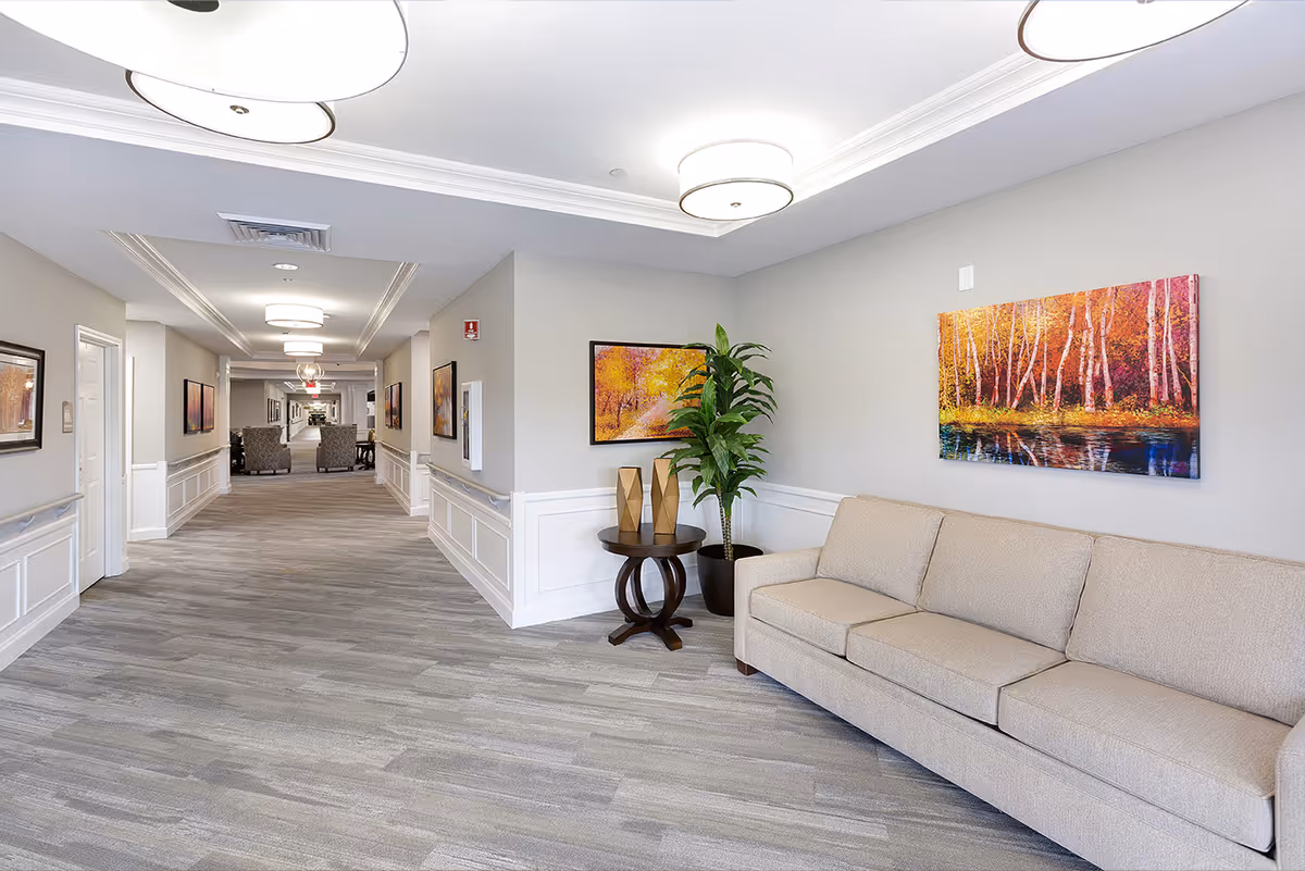 Well-lit senior living facility hallway with a beige sofa, side table, potted plant, and colorful wall art.