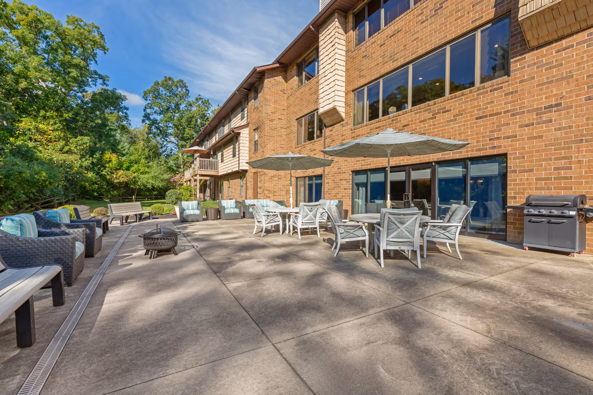 Outdoor patio area at Brookdale Gardens at Westlake featuring multiple seating arrangements with cushioned chairs, tables with umbrellas, a fire pit, and a barbecue grill next to a brick building under a clear blue sky.